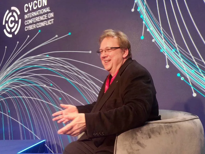 Andreas Lindenblatt sitzt lachend in einem Sessel auf der Bühne vor dem CyCon-Logo.
